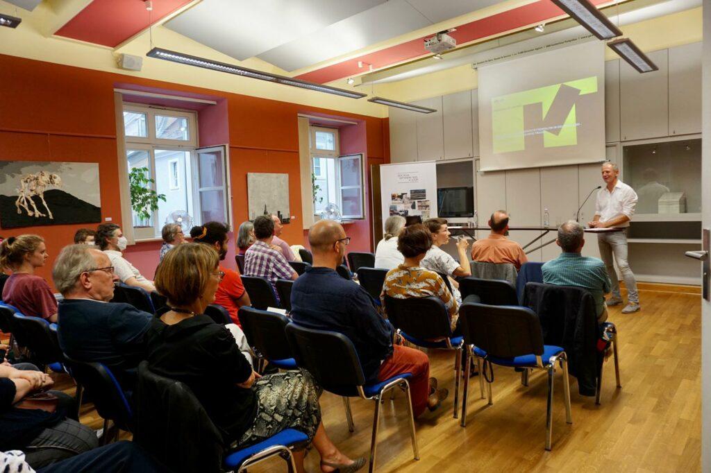 Das Bild zeigt das Publikum der Veranstaltung im Saal von hinten und Prof. Jörg Skriebeleit am Sprecherpult